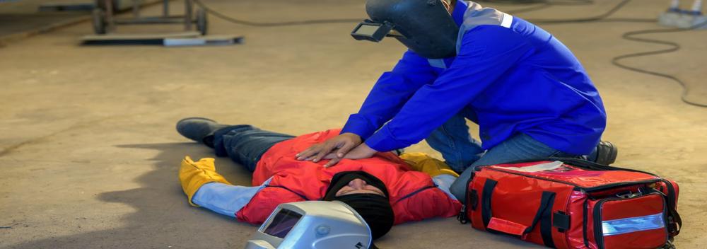 A welder performs CPR on another welder who lies on the ground unconscious after a workplace injury.